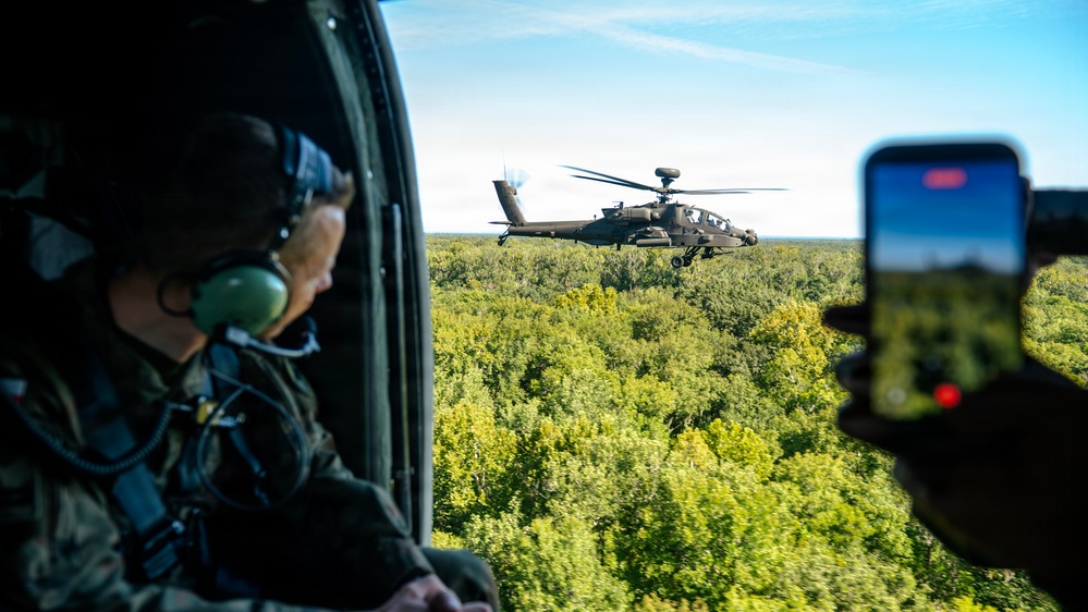 A military personnel wearing a flight helmet and green ear protection is seated inside a helicopter, observing an AH-64 Apache attack helicopter flying in close formation over a dense forest canopy. A second person in the foreground is recording the Apache helicopter using a smartphone, with the recording indicator visible on the screen. The scene appears to be a formation flight or demonstration exercise conducted in clear weather conditions.