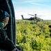 A military personnel wearing a flight helmet and green ear protection is seated inside a helicopter, observing an AH-64 Apache attack helicopter flying in close formation over a dense forest canopy. A second person in the foreground is recording the Apache helicopter using a smartphone, with the recording indicator visible on the screen. The scene appears to be a formation flight or demonstration exercise conducted in clear weather conditions.