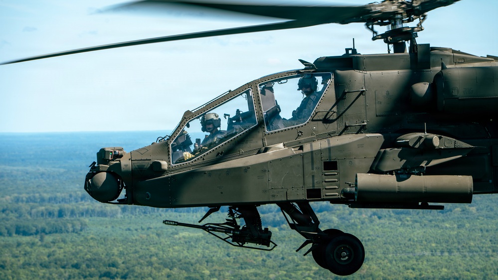 A military attack helicopter, consistent with an AH-64 Apache, is captured in flight over a densely forested landscape. Two crew members are visible in the tandem cockpit, wearing flight helmets and gear. The aircraft is painted in olive drab/dark green and appears fully equipped with sensors and weapons systems.