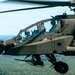 A military attack helicopter, consistent with an AH-64 Apache, is captured in flight over a densely forested landscape. Two crew members are visible in the tandem cockpit, wearing flight helmets and gear. The aircraft is painted in olive drab/dark green and appears fully equipped with sensors and weapons systems.