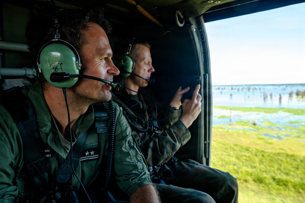 Two military personnel are seated inside a helicopter, observing a flooded landscape below. The individual in the foreground wears two-star rank insignia on their flight suit, suggesting a senior officer rank. The person in the background is using a tablet device, likely for reconnaissance or assessment purposes.
