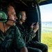 Two military personnel are seated inside a helicopter, observing a flooded landscape below. The individual in the foreground wears two-star rank insignia on their flight suit, suggesting a senior officer rank. The person in the background is using a tablet device, likely for reconnaissance or assessment purposes.