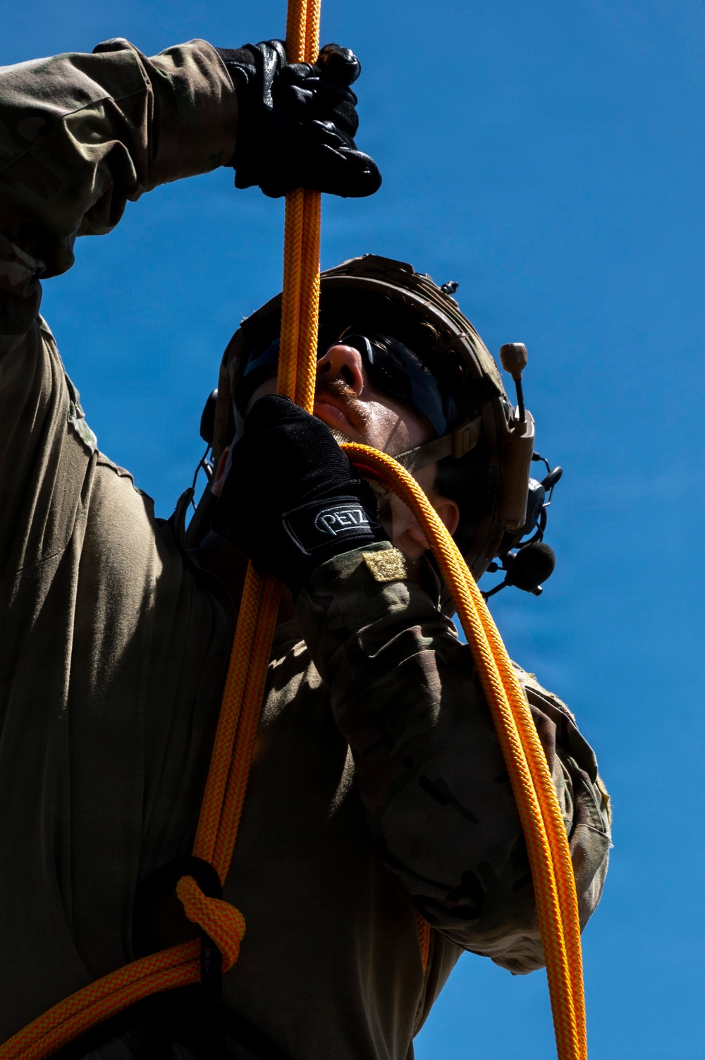 A person in military or tactical gear is handling an orange rope, viewed from a low angle against a clear blue sky. They are wearing a tactical helmet with communications equipment, dark sunglasses, and Petzl-branded gloves. The image captures an upward perspective of what appears to be a rappelling or rope-handling exercise.