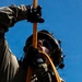 A person in military or tactical gear is handling an orange rope, viewed from a low angle against a clear blue sky. They are wearing a tactical helmet with communications equipment, dark sunglasses, and Petzl-branded gloves. The image captures an upward perspective of what appears to be a rappelling or rope-handling exercise.