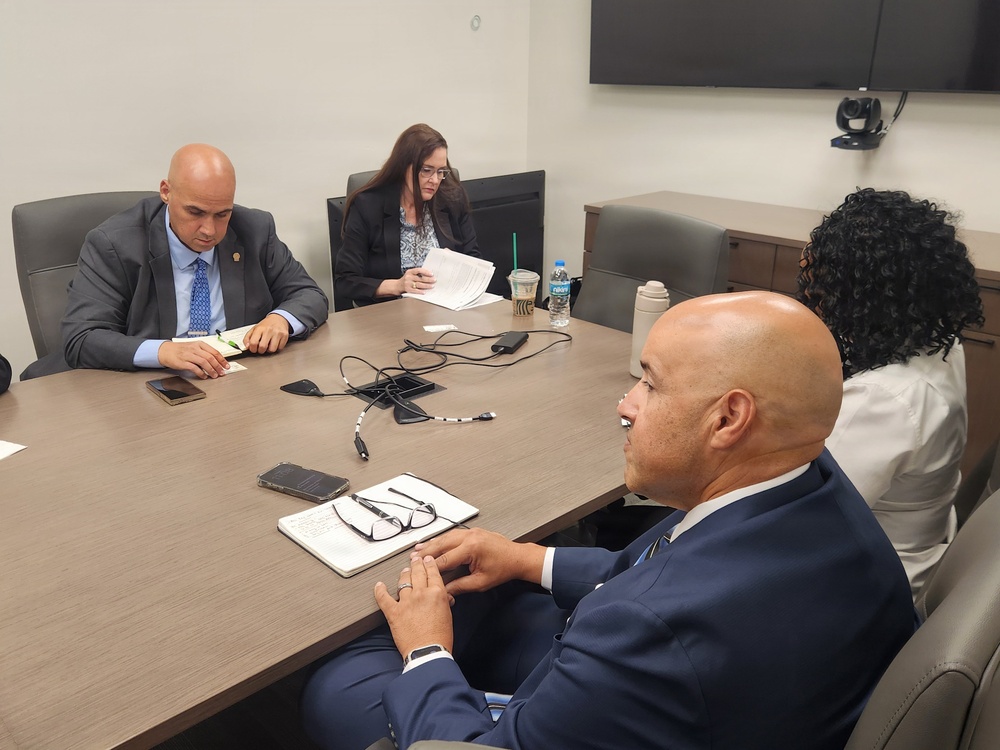 Four individuals are seated around a conference table in what appears to be a formal meeting or briefing setting. Two men in suits face each other across the table, while a woman reviews documents in the background and another woman sits partially visible in the foreground. The setting suggests a professional or governmental meeting environment.