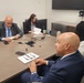 Four individuals are seated around a conference table in what appears to be a formal meeting or briefing setting. Two men in suits face each other across the table, while a woman reviews documents in the background and another woman sits partially visible in the foreground. The setting suggests a professional or governmental meeting environment.