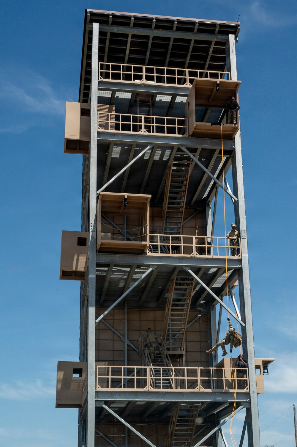 A multi-story military rappelling tower is shown with several personnel in military uniforms conducting rappelling training exercises. Multiple individuals are visible at different levels of the steel-framed structure, with at least one person actively rappelling down the exterior using a rope. The tower appears to be a purpose-built training facility with multiple platforms, staircases, and landing areas.