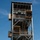 A multi-story military rappelling tower is shown with several personnel in military uniforms conducting rappelling training exercises. Multiple individuals are visible at different levels of the steel-framed structure, with at least one person actively rappelling down the exterior using a rope. The tower appears to be a purpose-built training facility with multiple platforms, staircases, and landing areas.