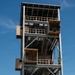 A multi-story military rappelling tower is shown with several personnel in military uniforms conducting rappelling training exercises. Multiple individuals are visible at different levels of the steel-framed structure, with at least one person actively rappelling down the exterior using a rope. The tower appears to be a purpose-built training facility with multiple platforms, staircases, and landing areas.