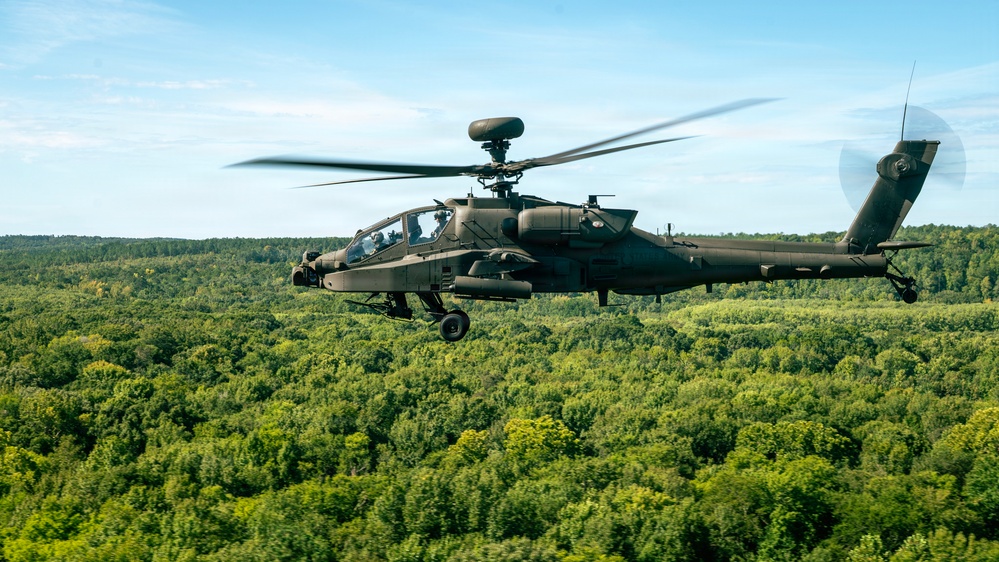 A military attack helicopter, identifiable as a Boeing AH-64 Apache, is captured in flight over a dense green forest canopy under a partly cloudy blue sky. The helicopter is painted in olive drab/dark green camouflage and appears to be in level forward flight. The main rotor blades show motion blur, indicating active flight.