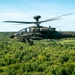 A military attack helicopter, identifiable as a Boeing AH-64 Apache, is captured in flight over a dense green forest canopy under a partly cloudy blue sky. The helicopter is painted in olive drab/dark green camouflage and appears to be in level forward flight. The main rotor blades show motion blur, indicating active flight.