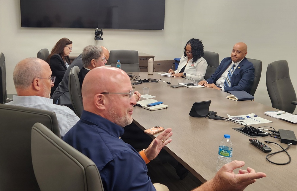 A professional meeting is taking place in a modern conference room with approximately six participants seated around a large wooden table. One man in a navy blue shirt appears to be actively speaking or presenting, with his hands gesturing. The group includes both men and women in business attire, with documents, tablets, and water bottles visible on the table.