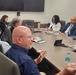 A professional meeting is taking place in a modern conference room with approximately six participants seated around a large wooden table. One man in a navy blue shirt appears to be actively speaking or presenting, with his hands gesturing. The group includes both men and women in business attire, with documents, tablets, and water bottles visible on the table.