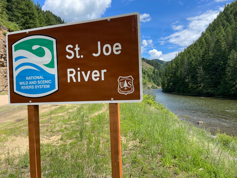 A brown informational sign identifies the St. Joe River as part of the National Wild and Scenic Rivers System, managed by the US Forest Service (Department of Agriculture). The sign is mounted on two wooden posts beside the river, which flows through a forested mountain canyon. The scene is photographed under partly cloudy skies with lush green vegetation visible throughout.