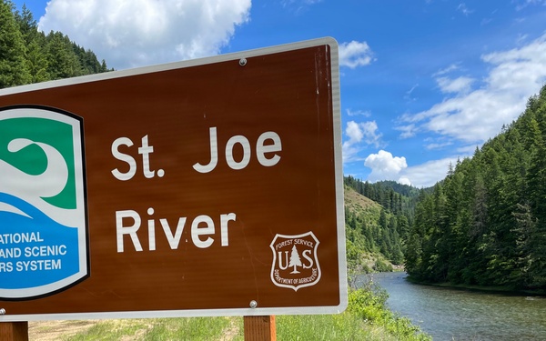 A brown informational sign identifies the St. Joe River as part of the National Wild and Scenic Rivers System, managed by the US Forest Service (Department of Agriculture). The sign is mounted on two wooden posts beside the river, which flows through a forested mountain canyon. The scene is photographed under partly cloudy skies with lush green vegetation visible throughout.