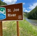 A brown informational sign identifies the St. Joe River as part of the National Wild and Scenic Rivers System, managed by the US Forest Service (Department of Agriculture). The sign is mounted on two wooden posts beside the river, which flows through a forested mountain canyon. The scene is photographed under partly cloudy skies with lush green vegetation visible throughout.