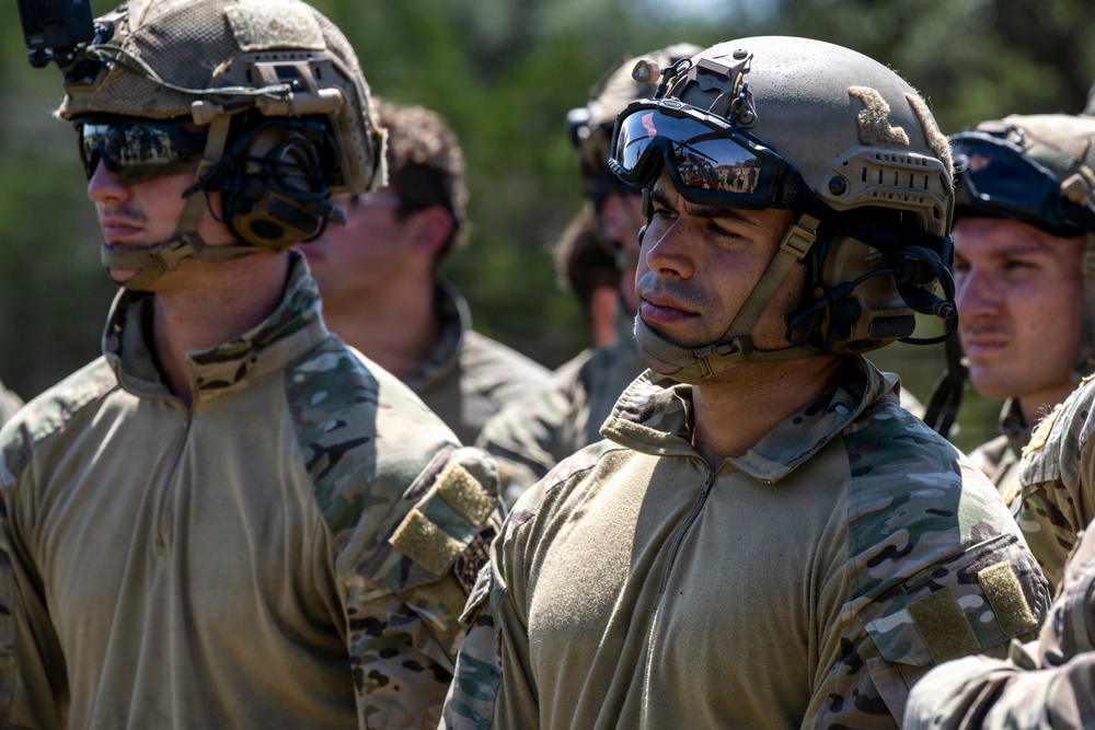 The image shows a group of military personnel in tactical gear standing in formation, likely during a training exercise or briefing. They are wearing multicam/OCP camouflage uniforms, tactical helmets with mounted accessories, and ballistic eyewear. The soldiers appear attentive and focused, suggesting they are receiving instructions or awaiting orders.