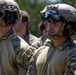 The image shows a group of military personnel in tactical gear standing in formation, likely during a training exercise or briefing. They are wearing multicam/OCP camouflage uniforms, tactical helmets with mounted accessories, and ballistic eyewear. The soldiers appear attentive and focused, suggesting they are receiving instructions or awaiting orders.