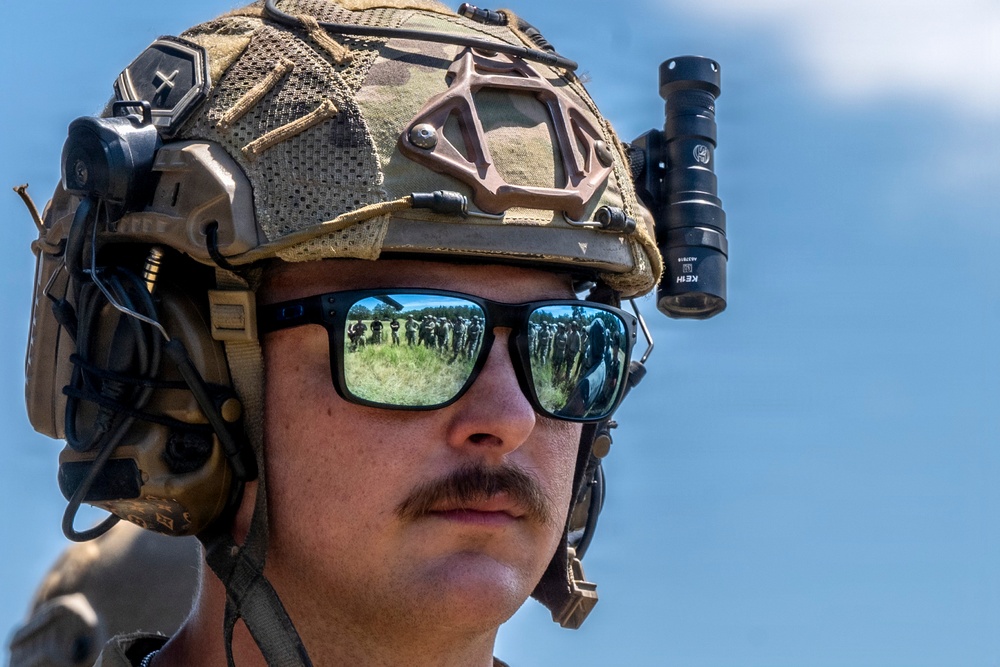 A close-up portrait of a military personnel wearing full tactical gear including a multicam FAST helmet with mounted flashlight and communication headset. The subject is wearing mirrored sunglasses that reflect a group of soldiers standing in a field. The image is a high-quality, sharp photograph taken outdoors under a clear blue sky.