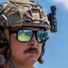 A close-up portrait of a military personnel wearing full tactical gear including a multicam FAST helmet with mounted flashlight and communication headset. The subject is wearing mirrored sunglasses that reflect a group of soldiers standing in a field. The image is a high-quality, sharp photograph taken outdoors under a clear blue sky.