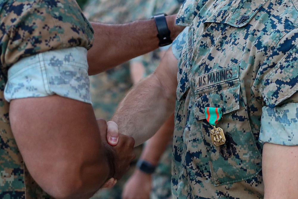 Two U.S. Marines in MARPAT digital camouflage uniforms are engaged in a handshake, suggesting a formal recognition or award ceremony. One Marine is wearing a medal with a teal/green and red/orange striped ribbon and a gold anchor-emblazoned pendant, consistent with a Navy achievement or commendation medal. The 'U.S. MARINES' chest tape and sergeant rank insignia are visible on one uniform.