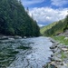 A clear, fast-moving river flows through a forested mountain valley flanked by dense coniferous trees on both sides. The foreground shows a rocky shoreline with green grass, while the river curves gently into the distance beneath a partly cloudy blue sky. The scene suggests a remote, wilderness setting likely in the Pacific Northwest or Northern Rocky Mountain region.