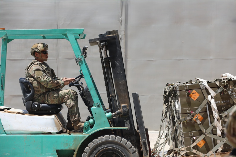 A military service member in full camouflage uniform and helmet is operating a teal/green forklift in what appears to be a logistics or supply area. The forklift is positioned near a palletized load of olive-drab military ammunition or supply crates secured with cargo netting and straps. Orange hazard warning diamonds are visible on the crates, indicating the presence of dangerous or explosive materials.