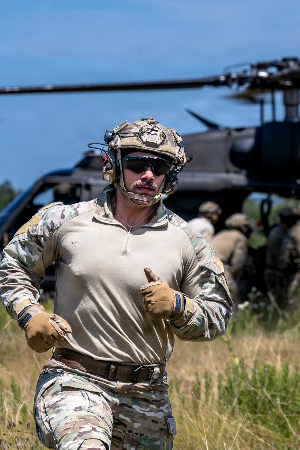 A military operator in full tactical gear is running away from a helicopter that has landed in a grassy field. Additional personnel are visible in the background near the aircraft, suggesting a helicopter insertion or extraction operation. The image captures an active field exercise or operational deployment under clear daylight conditions.
