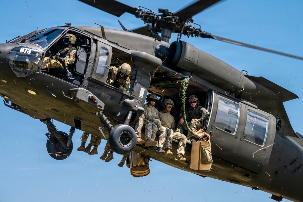 A military UH-60 Black Hawk helicopter (tail number 673) is captured in flight against a clear blue sky. Multiple soldiers in full combat gear are visible both inside the cabin and hanging from the exterior of the aircraft. The image appears to document a military training or operational insertion exercise.