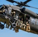 A military UH-60 Black Hawk helicopter (tail number 673) is captured in flight against a clear blue sky. Multiple soldiers in full combat gear are visible both inside the cabin and hanging from the exterior of the aircraft. The image appears to document a military training or operational insertion exercise.