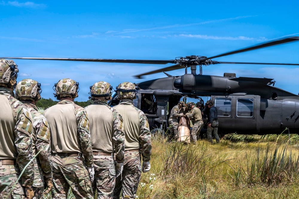 A group of military personnel in camouflage uniforms and tactical helmets are gathered near a black UH-60 Black Hawk helicopter landed in an open grassy field. Several soldiers are actively loading or unloading what appears to be a large orange/tan bag or casualty litter into the aircraft. The scene appears to depict a military training exercise or operational insertion/extraction scenario.