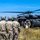 TACP Airmen master vertical infiltration A group of military personnel in camouflage uniforms and tactical helmets are gathered near a black UH-60 Black Hawk helicopter landed in an open grassy field. Several soldiers are actively loading or unloading what appears to be a large orange/tan bag or casualty litter into the aircraft. The scene appears to depict a military training exercise or operational insertion/extraction scenario.