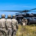 A group of military personnel in camouflage uniforms and tactical helmets are gathered near a black UH-60 Black Hawk helicopter landed in an open grassy field. Several soldiers are actively loading or unloading what appears to be a large orange/tan bag or casualty litter into the aircraft. The scene appears to depict a military training exercise or operational insertion/extraction scenario.