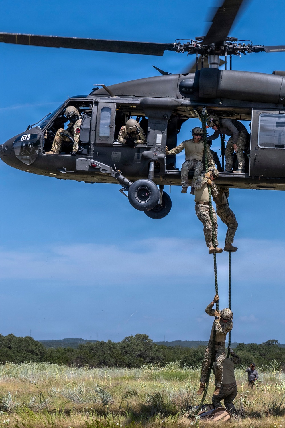Military personnel are conducting a fast-rope insertion/extraction (FRIES) training exercise from a UH-60 Black Hawk helicopter numbered 673. Multiple soldiers in camouflage uniforms and tactical gear are descending a thick rope from the hovering aircraft to the ground below. The exercise takes place over an open grassy field with trees visible in the background under clear blue skies.