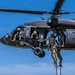 Military personnel are conducting a fast-rope insertion/extraction (FRIES) training exercise from a UH-60 Black Hawk helicopter numbered 673. Multiple soldiers in camouflage uniforms and tactical gear are descending a thick rope from the hovering aircraft to the ground below. The exercise takes place over an open grassy field with trees visible in the background under clear blue skies.