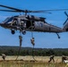 A United States Army UH-60 Black Hawk helicopter hovers low over an open field while soldiers conduct fast-rope insertion/extraction (FRIES) training. Multiple soldiers are actively rappelling down ropes from the aircraft while others have already landed and are moving away on the ground. The setting appears to be a military training area with open grassland and tree lines in the background.