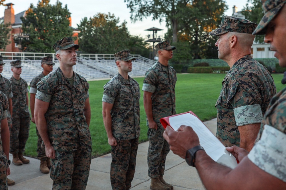 A group of U.S. Marines in MARPAT woodland camouflage uniforms are standing at attention during what appears to be a formal military ceremony or formation. A senior officer or NCO is addressing the assembled Marines while another individual in the foreground holds a red clipboard with documents. The setting appears to be a well-maintained outdoor parade ground or courtyard, likely on a military installation.