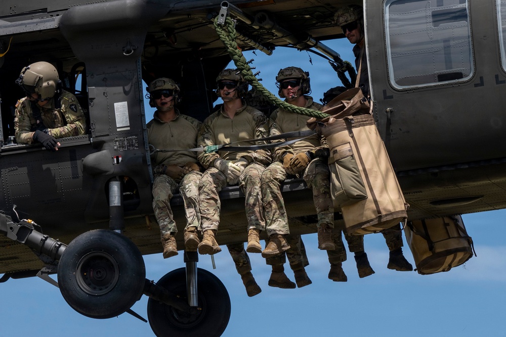 Military personnel are seated in the open doorway of a dark-colored helicopter in flight, with their legs dangling outside the aircraft. Several soldiers wearing camouflage uniforms, tactical helmets, and sunglasses are visible, along with tan-colored equipment bags secured to the exterior. A green rope is visible inside the cabin, suggesting preparation for a rappel or fast-rope insertion operation.