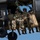 TACP Airmen master vertical infiltration Military personnel are seated in the open doorway of a dark-colored helicopter in flight, with their legs dangling outside the aircraft. Several soldiers wearing camouflage uniforms, tactical helmets, and sunglasses are visible, along with tan-colored equipment bags secured to the exterior. A green rope is visible inside the cabin, suggesting preparation for a rappel or fast-rope insertion operation.