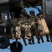 Military personnel are seated in the open doorway of a dark-colored helicopter in flight, with their legs dangling outside the aircraft. Several soldiers wearing camouflage uniforms, tactical helmets, and sunglasses are visible, along with tan-colored equipment bags secured to the exterior. A green rope is visible inside the cabin, suggesting preparation for a rappel or fast-rope insertion operation.