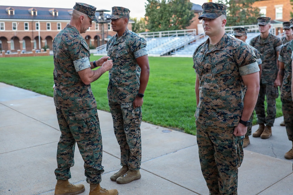A military ceremony is taking place outdoors on a paved area, where a senior Marine is pinning a decoration or device onto another Marine's uniform. Several other Marines in MARPAT camouflage uniforms stand in formation observing the ceremony. The setting appears to be a military installation with brick buildings and bleachers visible in the background.