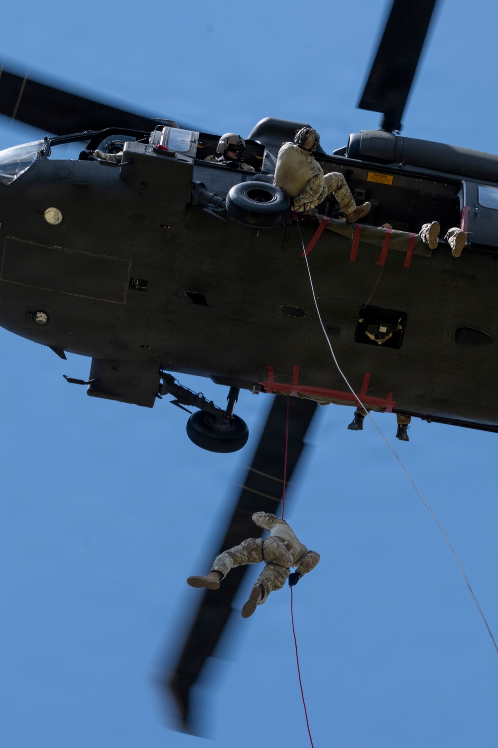 A military helicopter, appearing to be a UH-60 Black Hawk, is photographed from below against a clear blue sky. Multiple personnel in military camouflage uniforms are visible — one seated at the open door and at least one rappelling/fast-roping down from the aircraft on a rope. The image captures an active military fast-rope or rappel insertion training exercise.