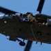 A military helicopter, appearing to be a UH-60 Black Hawk, is photographed from below against a clear blue sky. Multiple personnel in military camouflage uniforms are visible — one seated at the open door and at least one rappelling/fast-roping down from the aircraft on a rope. The image captures an active military fast-rope or rappel insertion training exercise.