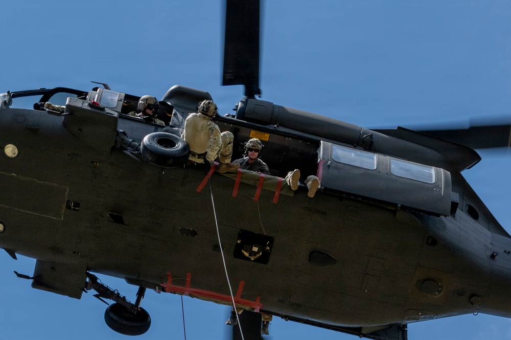 A military helicopter, viewed from below at an angle, is in flight with its side door open. Several personnel in military gear and helmets are visible inside the open cabin, with one individual sitting at the edge with legs dangling out. A rope or cable is visible hanging from the underside of the aircraft.