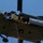 TACP Airmen master vertical infiltration A military helicopter, viewed from below at an angle, is in flight with its side door open. Several personnel in military gear and helmets are visible inside the open cabin, with one individual sitting at the edge with legs dangling out. A rope or cable is visible hanging from the underside of the aircraft.