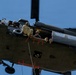 A military helicopter, viewed from below at an angle, is in flight with its side door open. Several personnel in military gear and helmets are visible inside the open cabin, with one individual sitting at the edge with legs dangling out. A rope or cable is visible hanging from the underside of the aircraft.