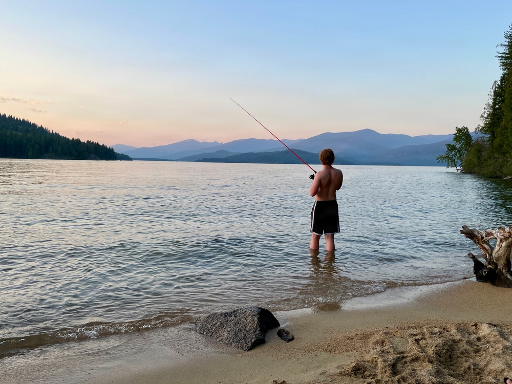 A shirtless young person stands ankle-deep in a calm lake at dusk, fishing with a red rod. The scenic backdrop features layered mountain ranges, forested shorelines, and a pastel sunset sky. A sandy beach with a large rock and driftwood frames the foreground.