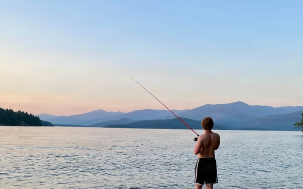 A shirtless young person stands ankle-deep in a calm lake at dusk, fishing with a red rod. The scenic backdrop features layered mountain ranges, forested shorelines, and a pastel sunset sky. A sandy beach with a large rock and driftwood frames the foreground.