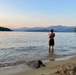 A shirtless young person stands ankle-deep in a calm lake at dusk, fishing with a red rod. The scenic backdrop features layered mountain ranges, forested shorelines, and a pastel sunset sky. A sandy beach with a large rock and driftwood frames the foreground.