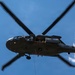 A military helicopter, consistent with a UH-60 Black Hawk, is photographed from below against a clear blue sky. A soldier in full combat gear is suspended beneath the aircraft on ropes, performing a fast-rope or rappelling operation. Red markings are visible on the underside of the helicopter, and at least one crew member is visible in the open door.